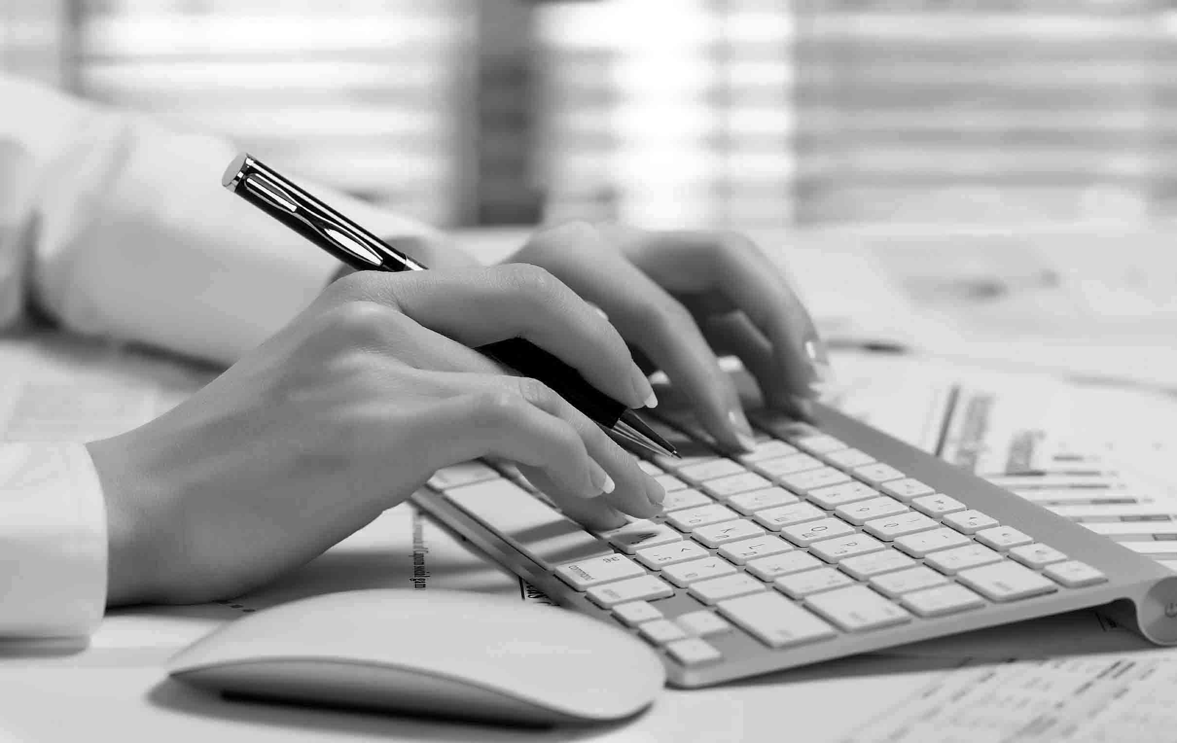female with pen in her right hand and typing into her keyboard